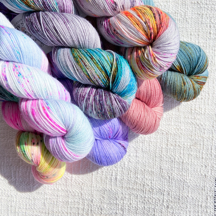 An image of seven skeins of La Bien Aimee sock yarn in a variety of colors against a canvas background.