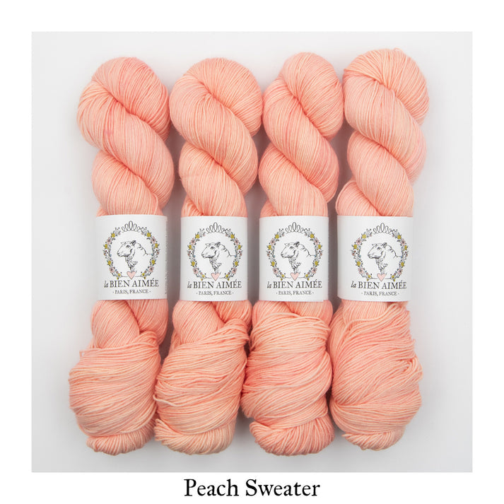 An image of four skeins of sock yarn in a peach pink shade with white ‘La Bien Aimee’ labels against a white background.