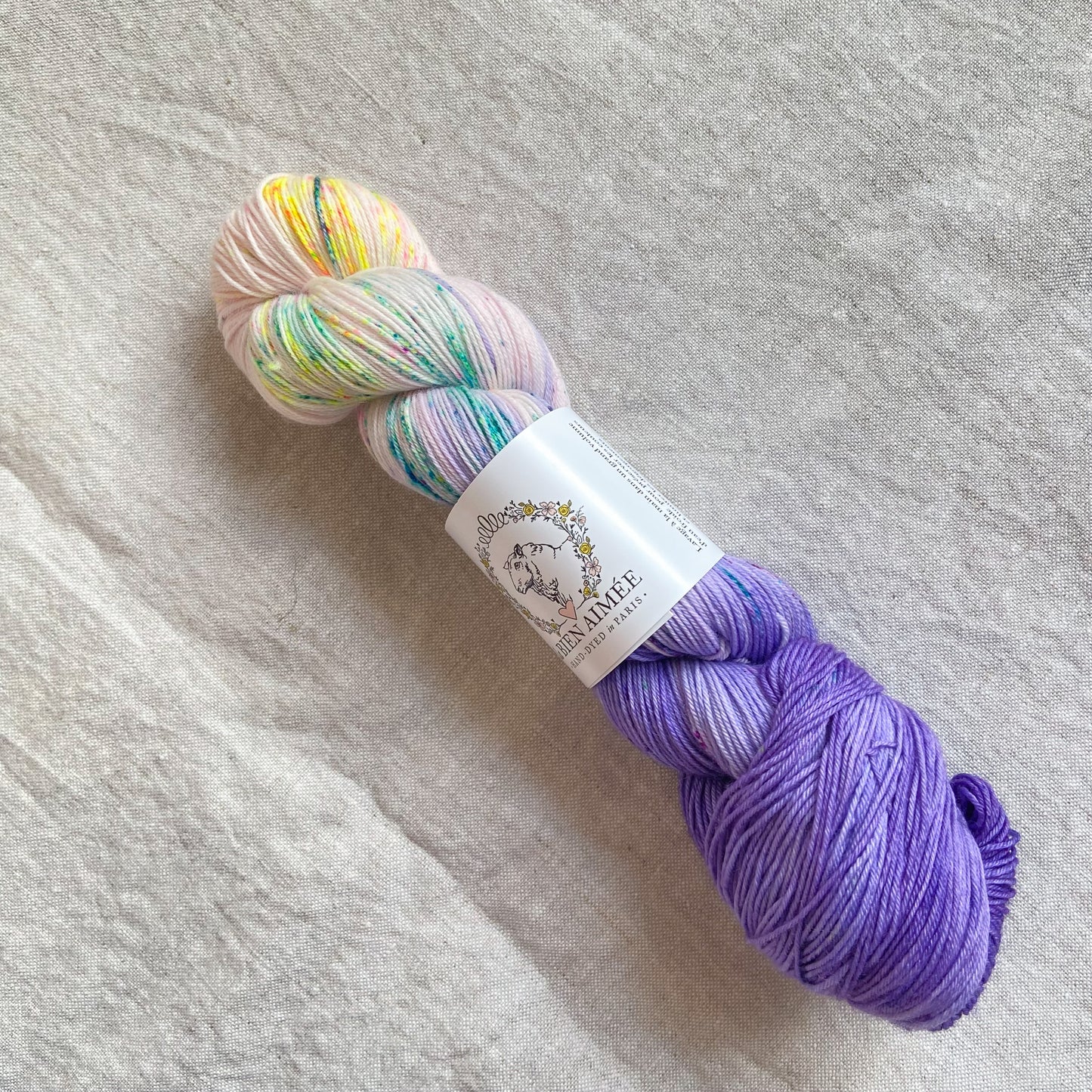 An image of a skein of sock yarn in a purple and cream base with speckles of blue and yellow, with a white ‘La Bien Aimee’ label against a canvas background.