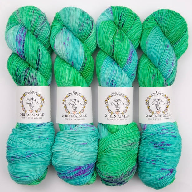 An image of four skeins of sock yarn in shades of aqua blue and green, with speckles of purple, with white ‘La Bien Aimee’ labels against a white background.