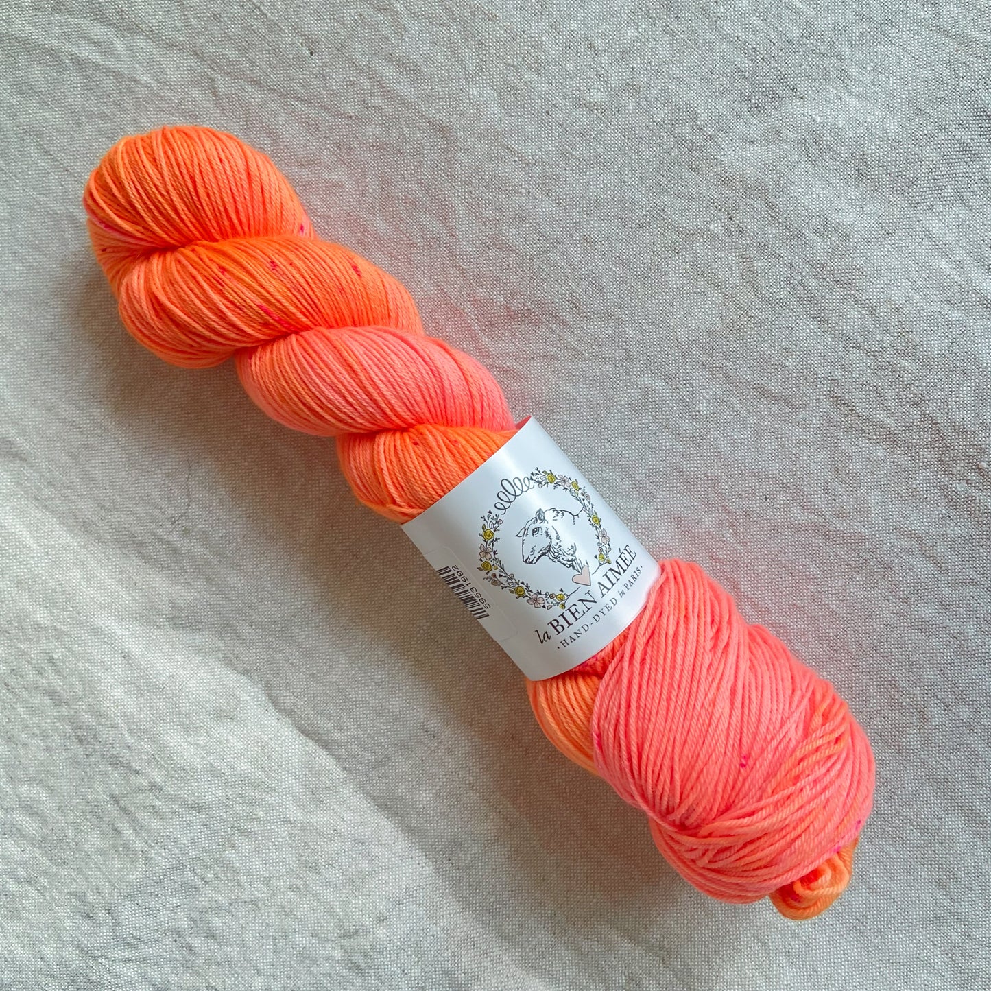 An image of a skein of sock yarn in neon shades of orange and pink, with a white ‘La Bien Aimee’ label against a canvas background.
