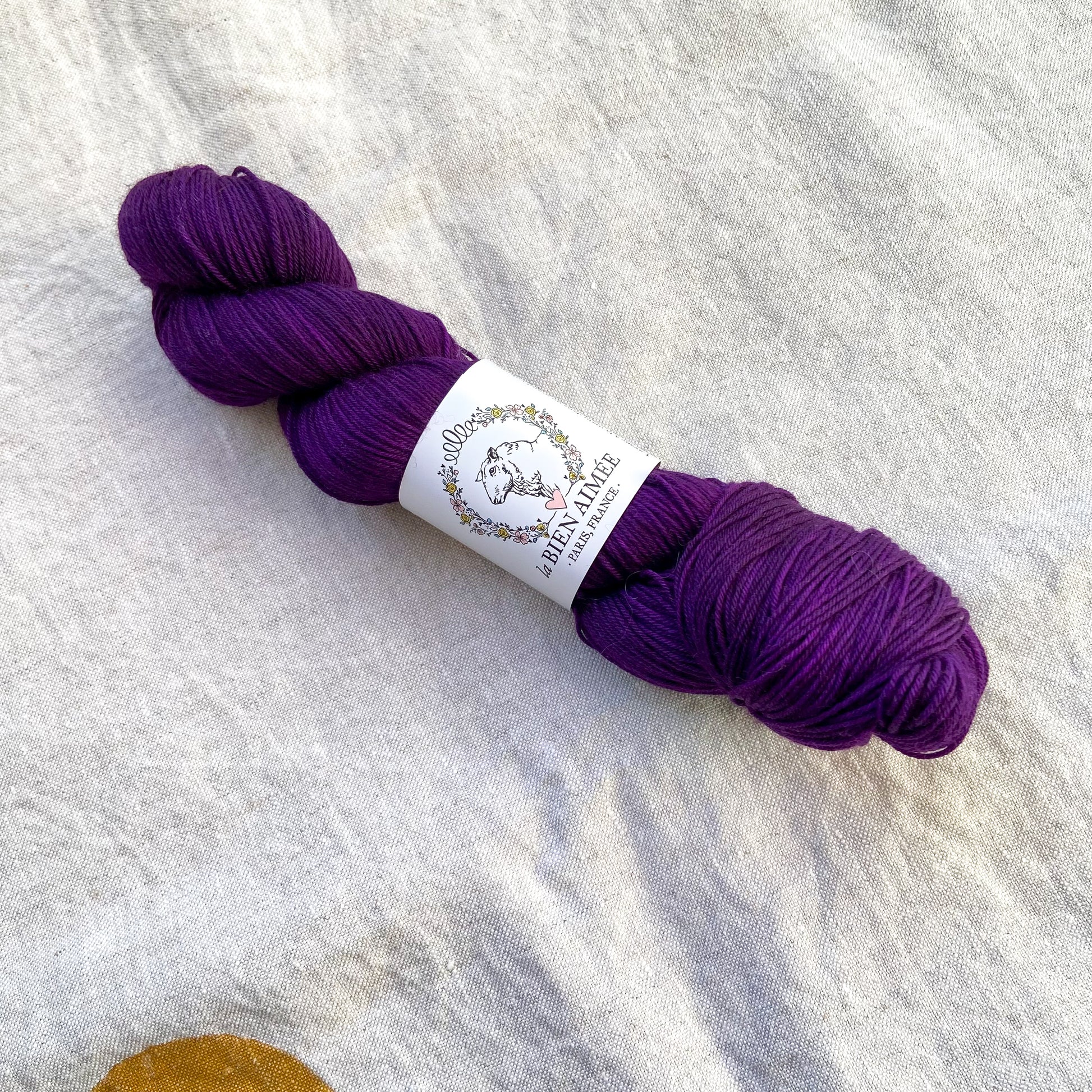 An image of a skein of sock yarn in a dark purple color, with a white ‘La Bien Aimee’ label against a canvas background.