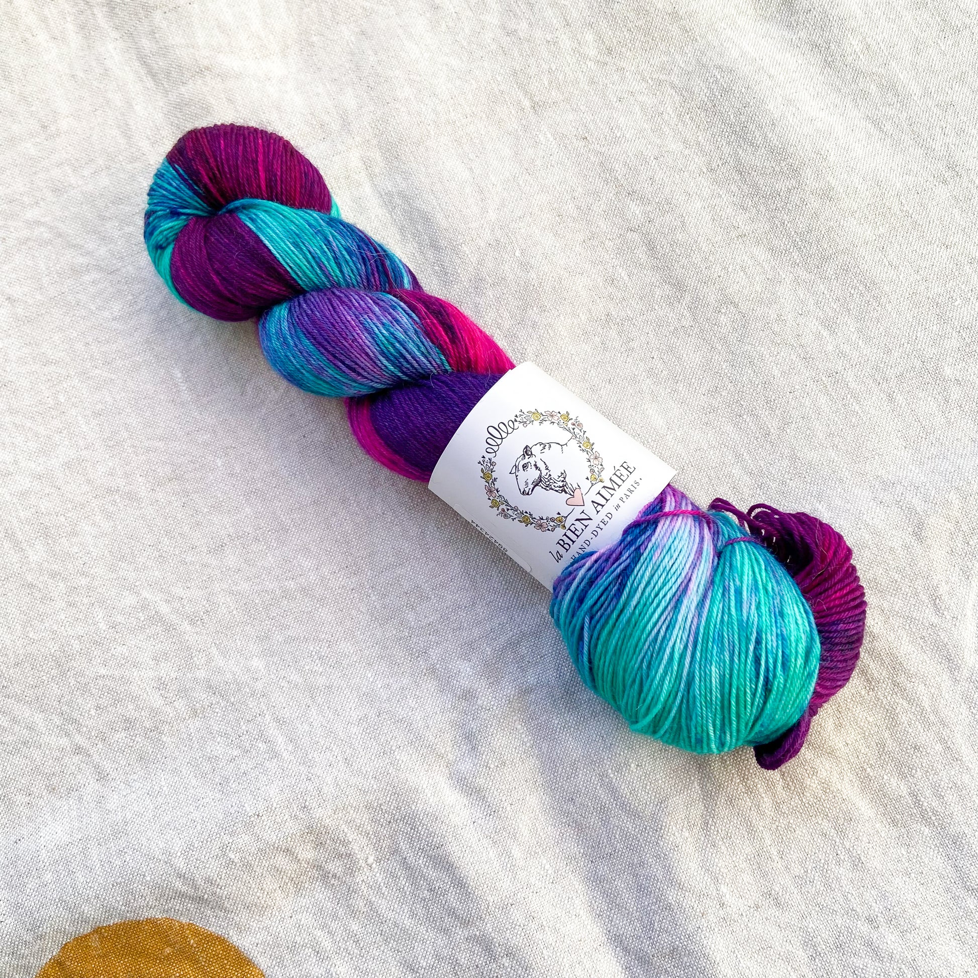 An image of a skein of sock yarn in shades of aqua blue, magenta, and purple, with a white ‘La Bien Aimee’ label against a canvas background.