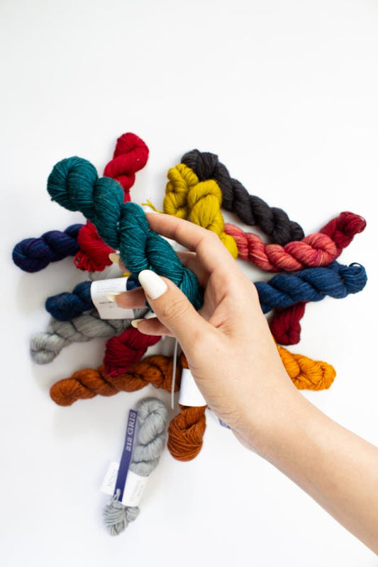 Hand holding a a mini skein of teal yarn, with several colorful mini skeins in the background, against a white background