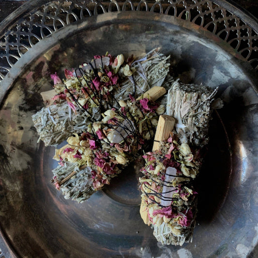 Two ritual bundles made of mugwort and palo santo, adorned with a crystal and herbs, displayed on a metal plate.