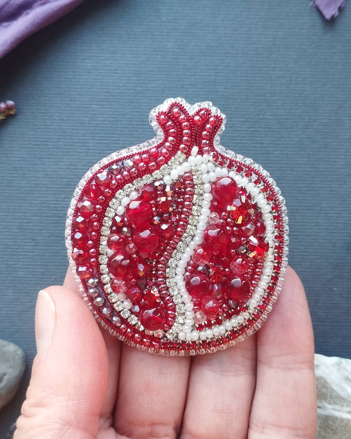 Hand holding a red and silver beaded pomegranate brooch on a textured surface with plants and stones.