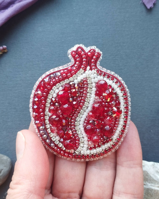 Hand holding a red and silver beaded pomegranate brooch on a textured surface with plants and stones.