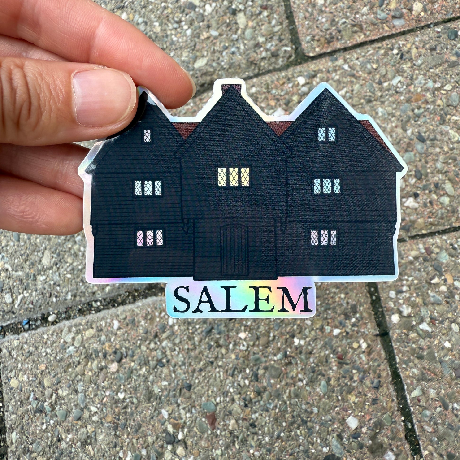 A holographic sticker featuring an illustration of the Witch House in Salem, Massachusetts. A hand is holding the sticker over a stone sidewalk. 