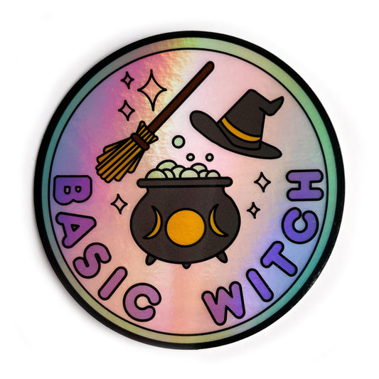 A circular holographic sticker. There is a mint green border around a pastel pink circle in the background. The bottom of the pin has bubble letters in purple that read "Basic Witch" above this there is a black cauldron with yellow moon phases full of bubbles. There is a broom above this and a black pointed hat to the right of the broom. Sparkles are around all of the objects.