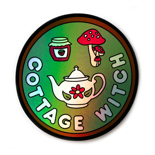 A circular holographic sticker. It has a brown border with a sage green background and the words "Cottage Witch" in light blue. Above the words are a teapot with a flower, a strawberry jam jar, and a red mushroom with white spots.
