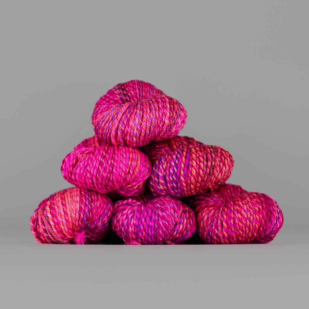 An image of skeins of Spincycle Dream State worsted weight yarn in a hot pink, purple, and maroon color gradient against a gray background.