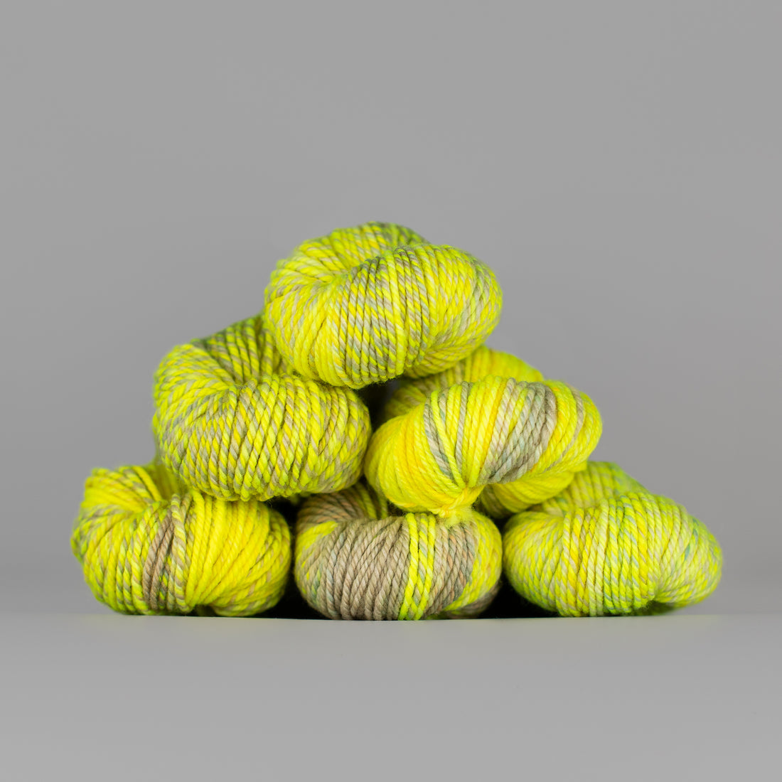 An image of skeins of Spincycle Dream State worsted weight yarn in a neon yellow and taupe color gradient against a gray background.