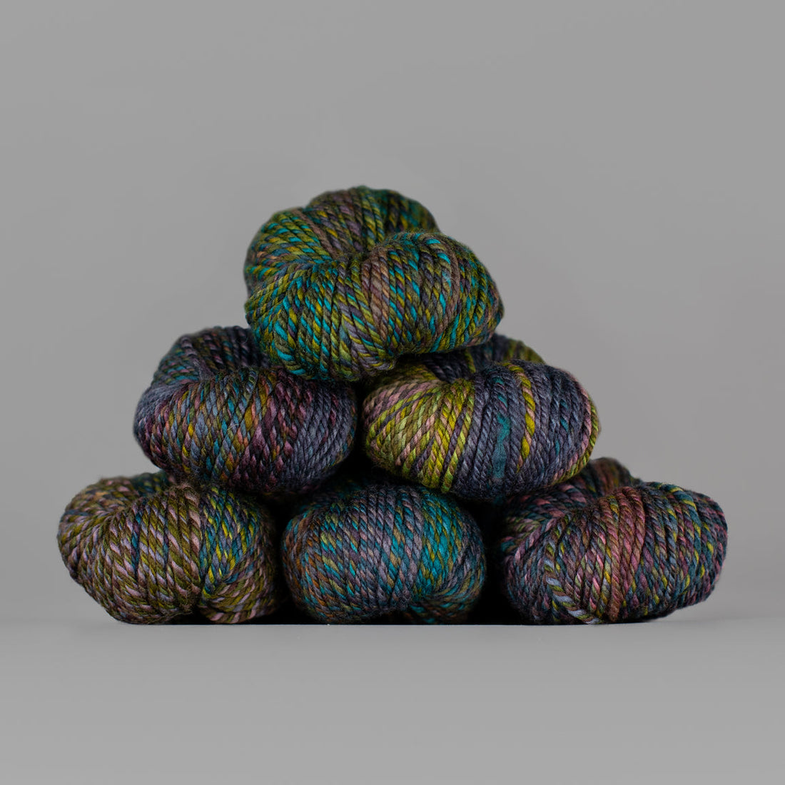 An image of skeins of Spincycle Dream State worsted weight yarn in a dark green, teal, purple, and brown color gradient against a gray background.