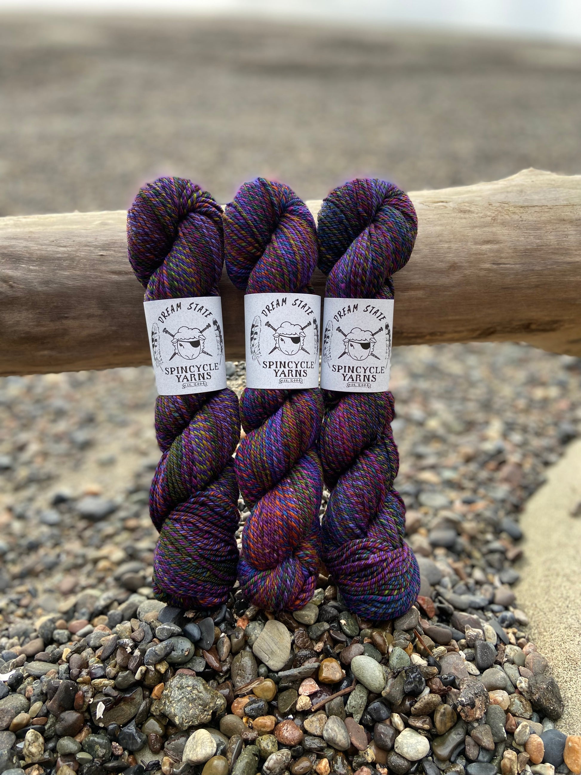 Three skeins of Spincycle Dream State worsted weight yarn in a blue, green, orange, and purple color gradient, with labels showing the brand 'Spincycle Yarns'  resting against a log on rocks.
