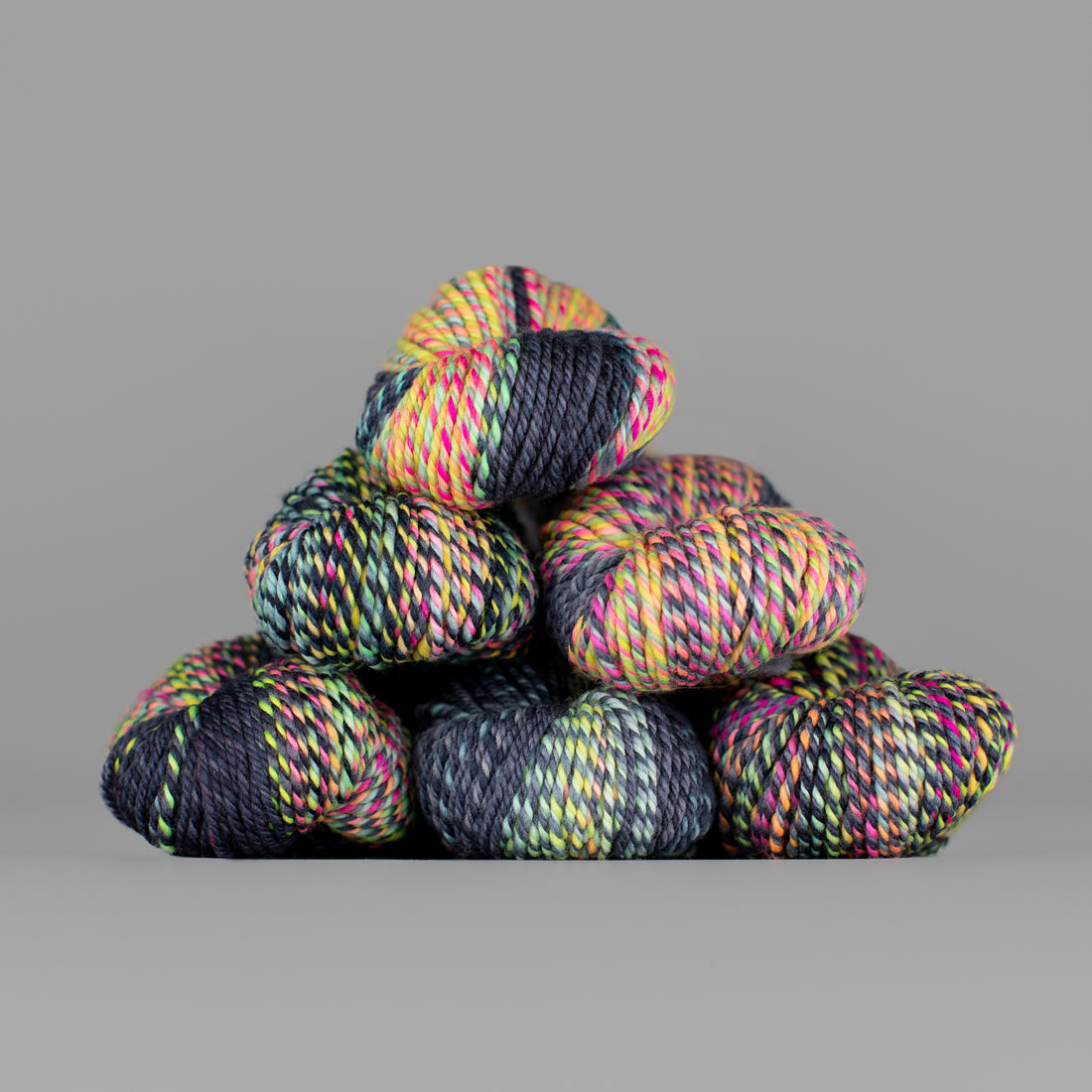 An image of skeins of Spincycle Dream State worsted weight yarn in a neon yellow, pink, and charcoal color gradient against a gray background.