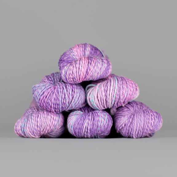 An image of skeins of Spincycle Dream State worsted weight yarn in a pale purple, pink, and blue color gradient against a gray background.