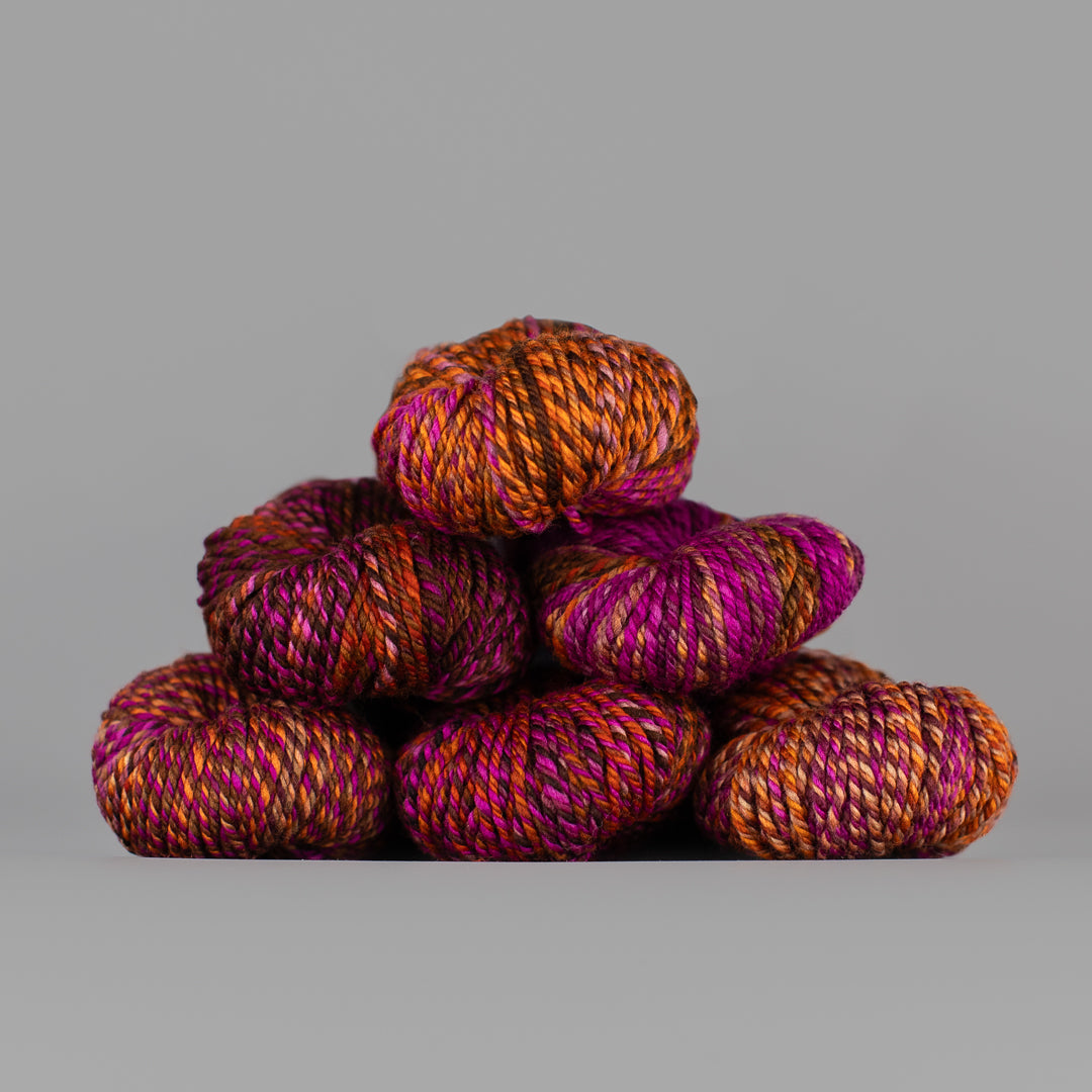 An image of skeins of Spincycle Dream State worsted weight yarn in a brown, orange, and pink color gradient against a gray background.
