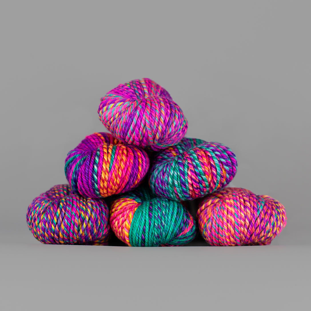 An image of skeins of Spincycle Dream State worsted weight yarn in a hot pink, orange, purple, and turquoise color gradient against a gray background.