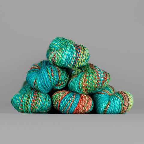 An image of skeins of Spincycle Dream State worsted weight yarn in an aqua blue, green, and red orange color gradient against a gray background.