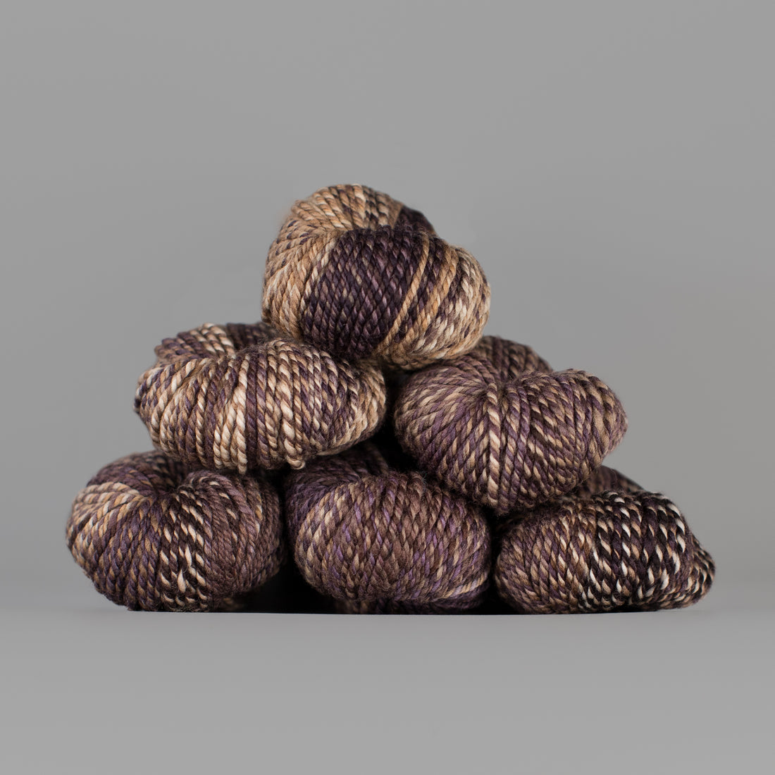 An image of skeins of Spincycle Dream State worsted weight yarn in a tan, mauve, and chocolate brown color gradient against a gray background.