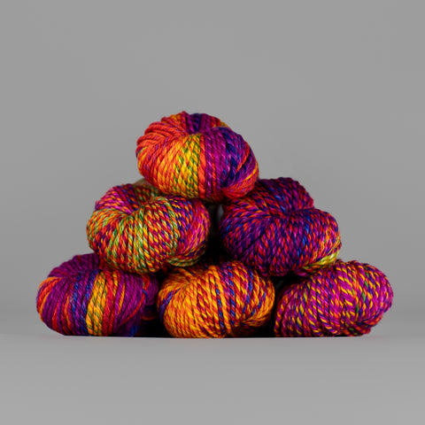 An image of skeins of Spincycle Dream State worsted weight yarn in a vibrant orange, magenta, and purple color gradient against a gray background.