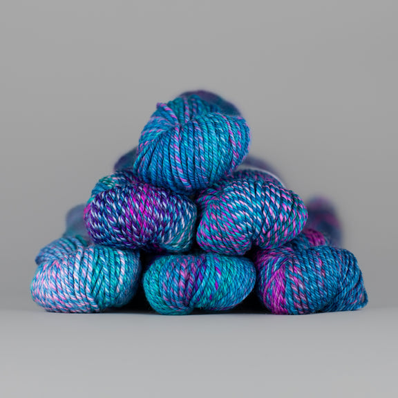 An image of skeins of Spincycle Dream State worsted weight yarn in an ocean blue, purple, and pink color gradient against a gray background.