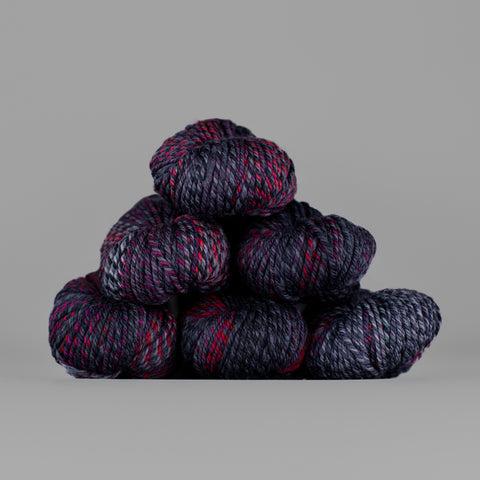 An image of skeins of Spincycle Dream State worsted weight yarn in a deep purple, magenta, and charcoal color gradient against a gray background.