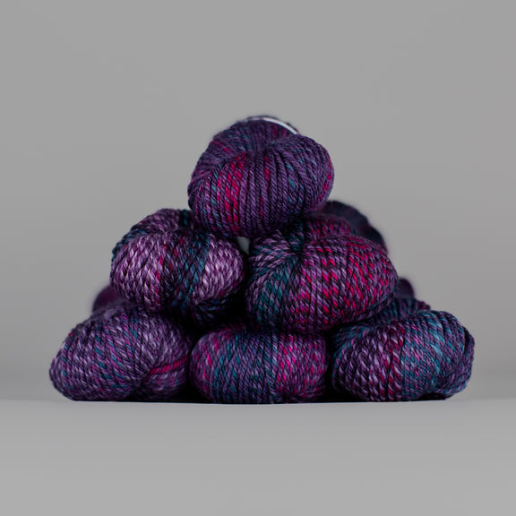 An image of skeins of Spincycle Dream State worsted weight yarn in a deep purple, magenta, and blue color gradient against a gray background.