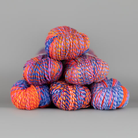 An image of skeins of Spincycle Dream State worsted weight yarn in a bright orange and purple color gradient against a gray background.