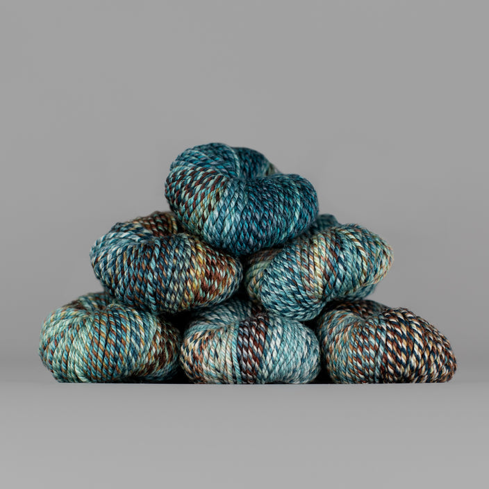 An image of skeins of Spincycle Dream State worsted weight yarn in a teal, gray blue, and brown color gradient against a gray background.