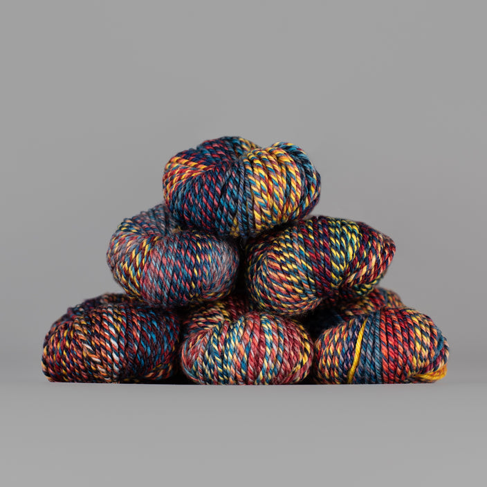 An image of skeins of Spincycle Dream State worsted weight yarn in a blue, red, yellow, and charcoal color gradient against a gray background.