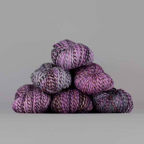 An image of skeins of Spincycle Dream State worsted weight yarn in a purple and gray color gradient against a gray background.