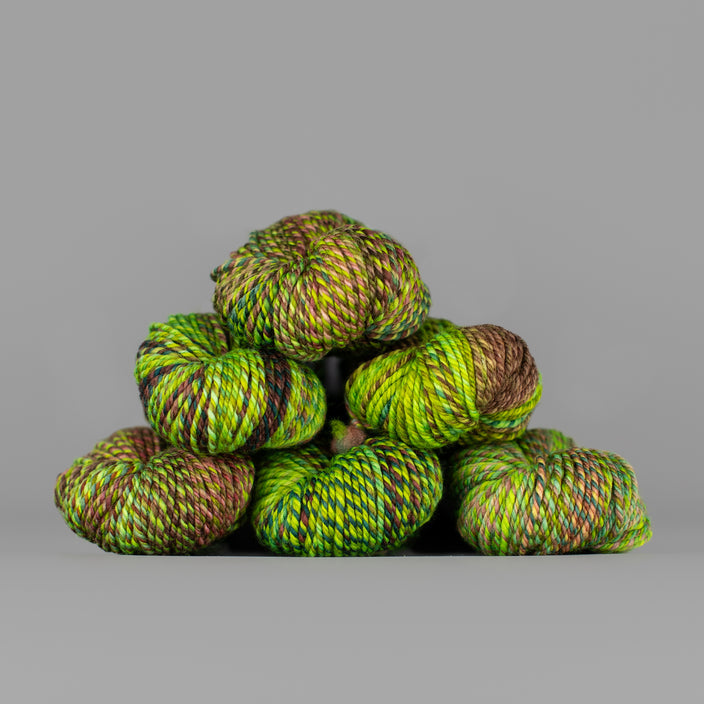An image of skeins of Spincycle Dream State worsted weight yarn in a neon green, forest green, and brown color gradient against a gray background.
