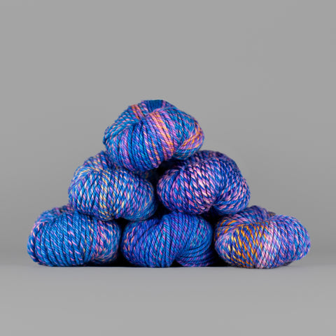 An image of skeins of Spincycle Dream State worsted weight yarn in an electric blue, peach pink, and purple color gradient against a gray background.