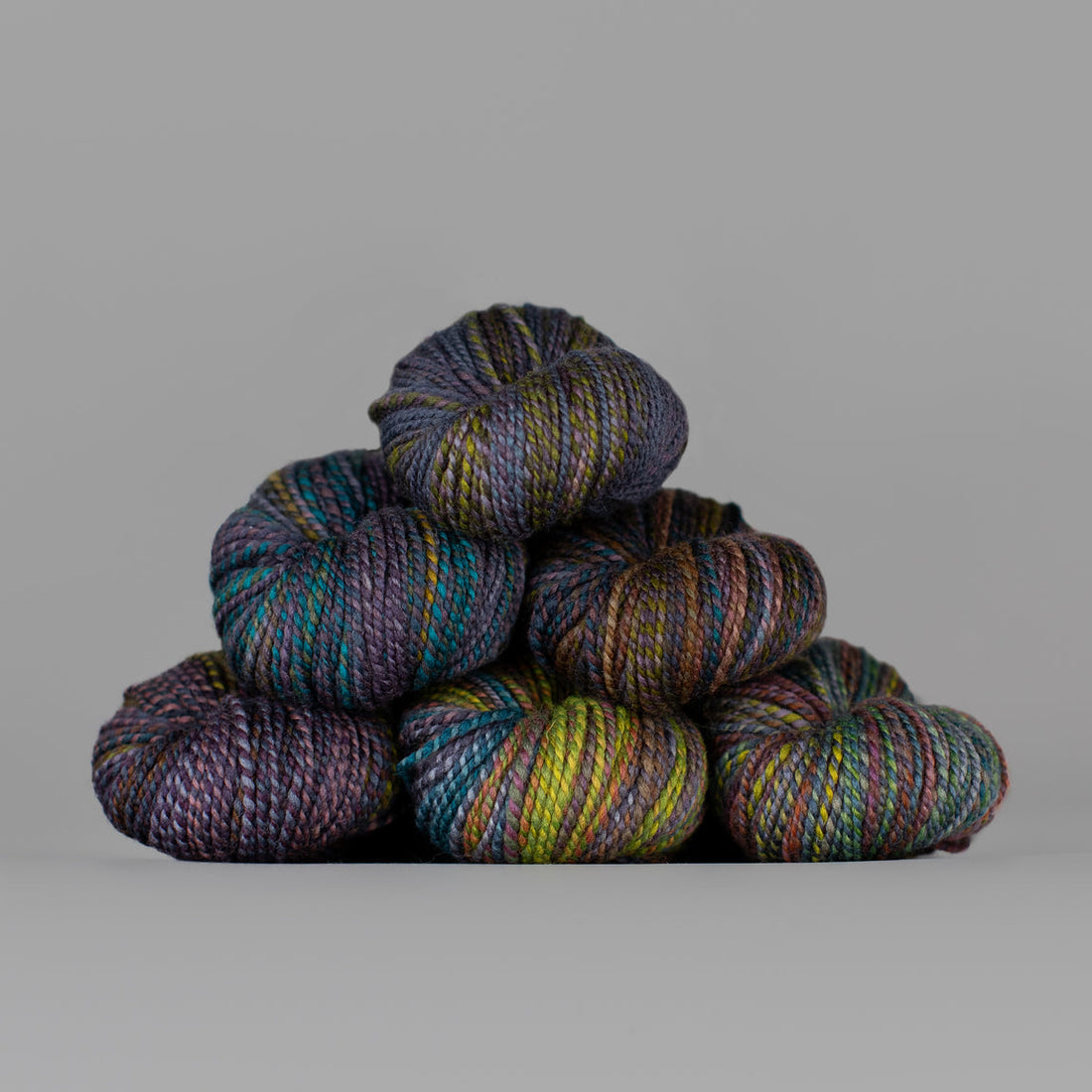 Skeins of Spincycle Dyed in the Wool sport weight yarn in a yellow green, olive green, teal, purple, and orange color gradient against a gray background.