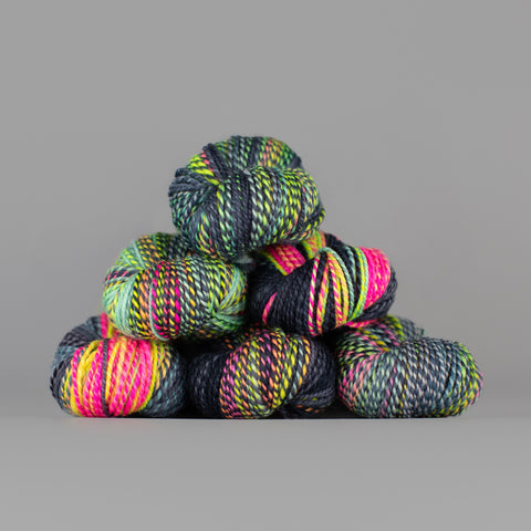 Skeins of Spincycle Dyed in the Wool sport weight yarn in a neon pink, yellow, green, and charcoal color gradient against a gray background.