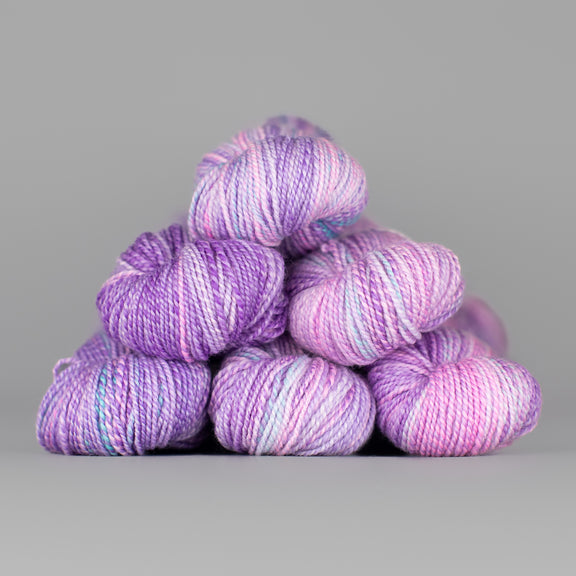 Skeins of Spincycle Dyed in the Wool sport weight yarn in a purple, pale pink, and blue color gradient against a gray background.