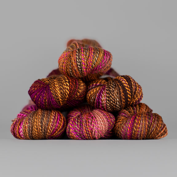 Skeins of Spincycle Dyed in the Wool sport weight yarn in a brown, orange, and pink color gradient against a gray background.