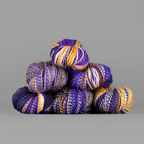 Skeins of Spincycle Dyed in the Wool sport weight yarn in an orange, purple, charcoal, and cream color gradient against a gray background.