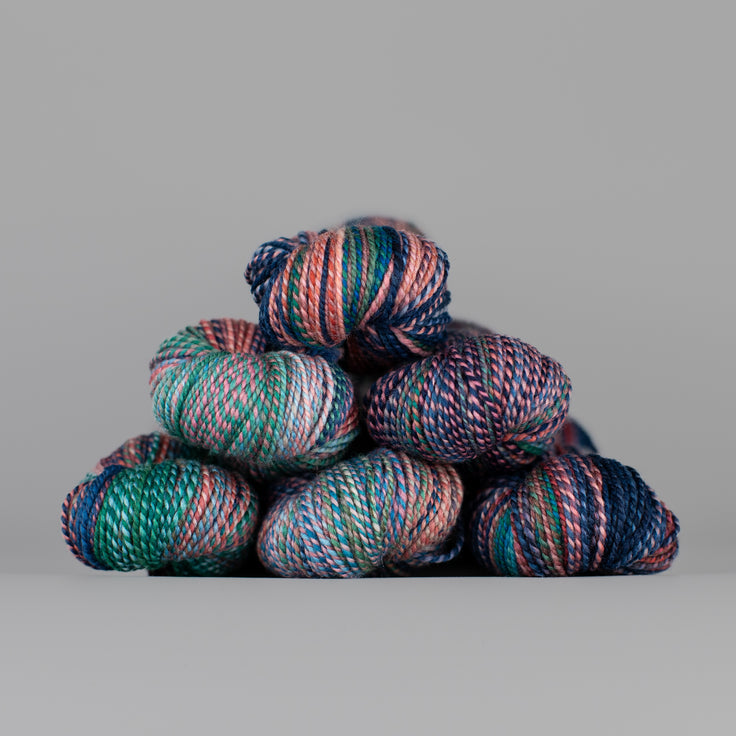 Skeins of Spincycle Dyed in the Wool sport weight yarn in a navy, teal, and light pink color gradient against a gray background.