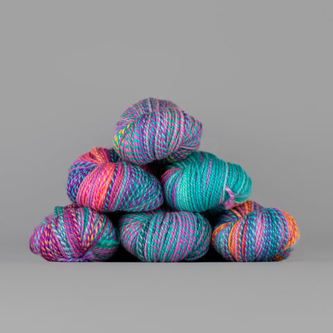 Skeins of Spincycle Dyed in the Wool sport weight yarn in an aqua blue, pink, orange, and purple color gradient against a gray background.