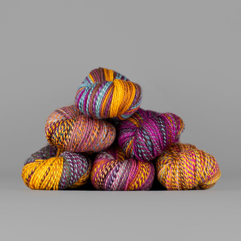 Skeins of Spincycle Dyed in the Wool sport weight yarn in a yellow gold, magenta, and blue gray color gradient against a gray background.