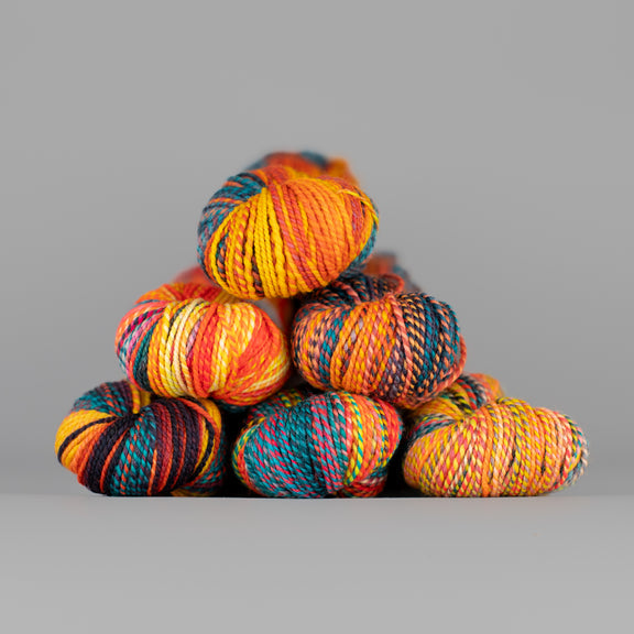 Skeins of Spincycle Dyed in the Wool sport weight yarn in a teal, red, orange, yellow, and purple color gradient against a gray background.
