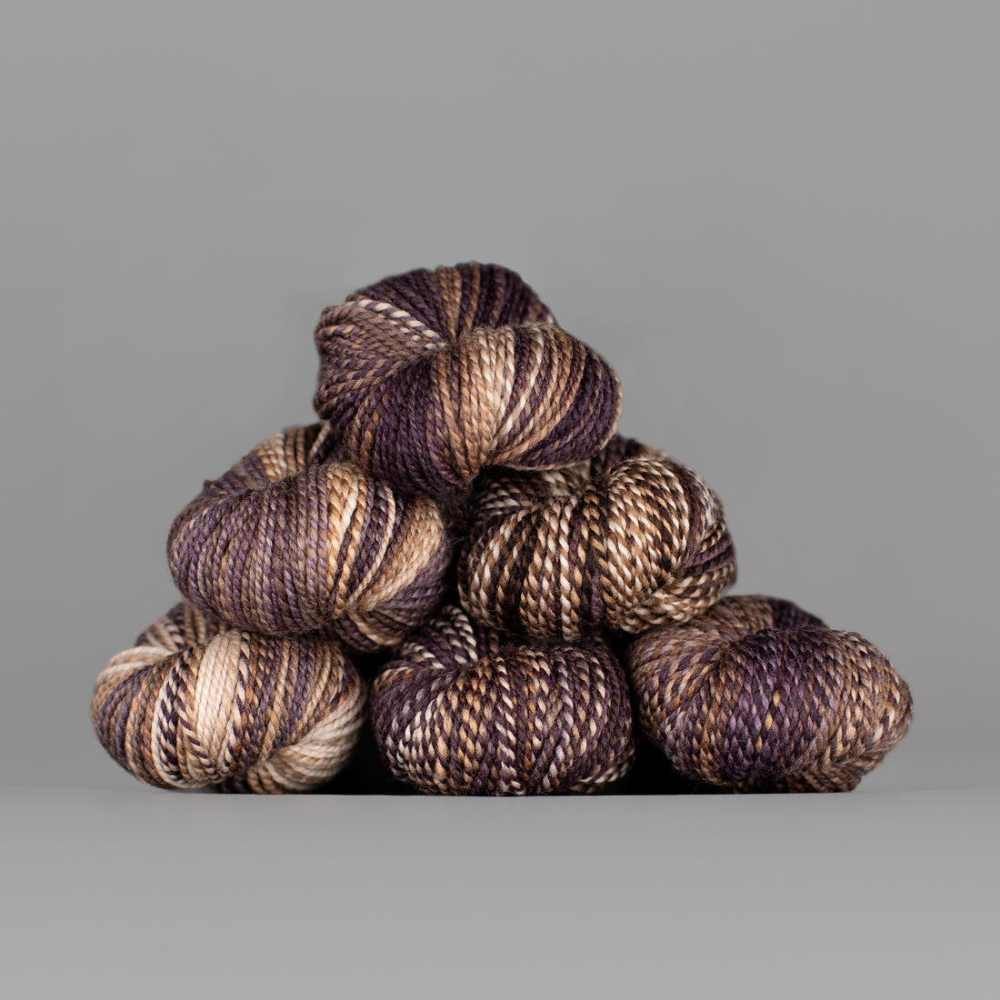 Skeins of Spincycle Dyed in the Wool sport weight yarn in a brown, cream, and mauve color gradient against a gray background.