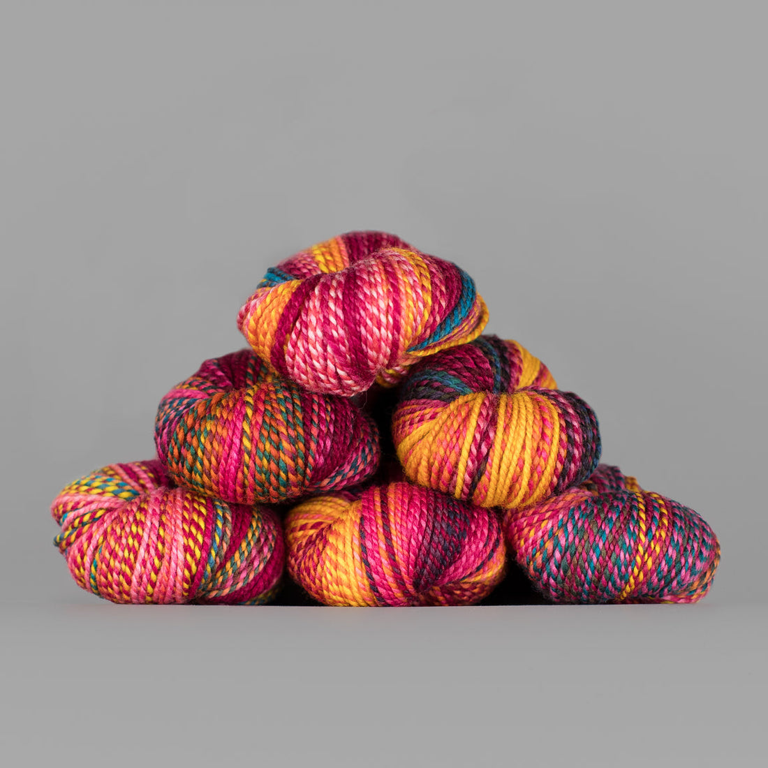 Skeins of Spincycle Dyed in the Wool sport weight yarn in a bright pink, yellow orange, teal, and charcoal color gradient against a gray background.