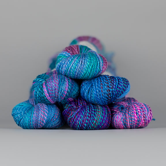 Skeins of Spincycle Dyed in the Wool sport weight yarn in an aqua, ocean blue, pink, and purple color gradient against a gray background.