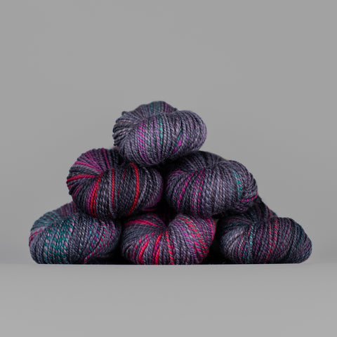 Skeins of Spincycle Dyed in the Wool sport weight yarn in a gray purple, teal, and hot pink color gradient against a gray background.
