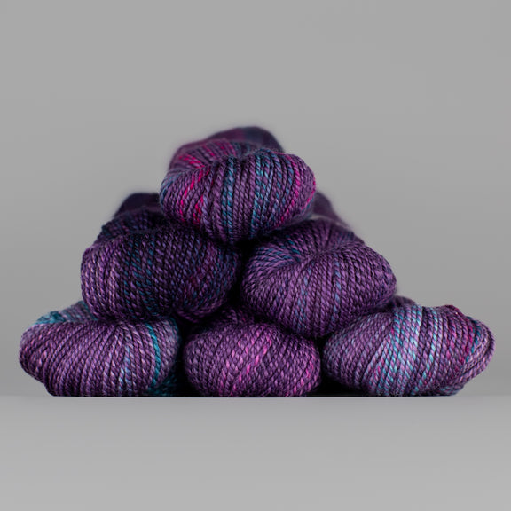 Skeins of Spincycle Dyed in the Wool sport weight yarn in a dark purple, blue, and pink color gradient against a gray background.