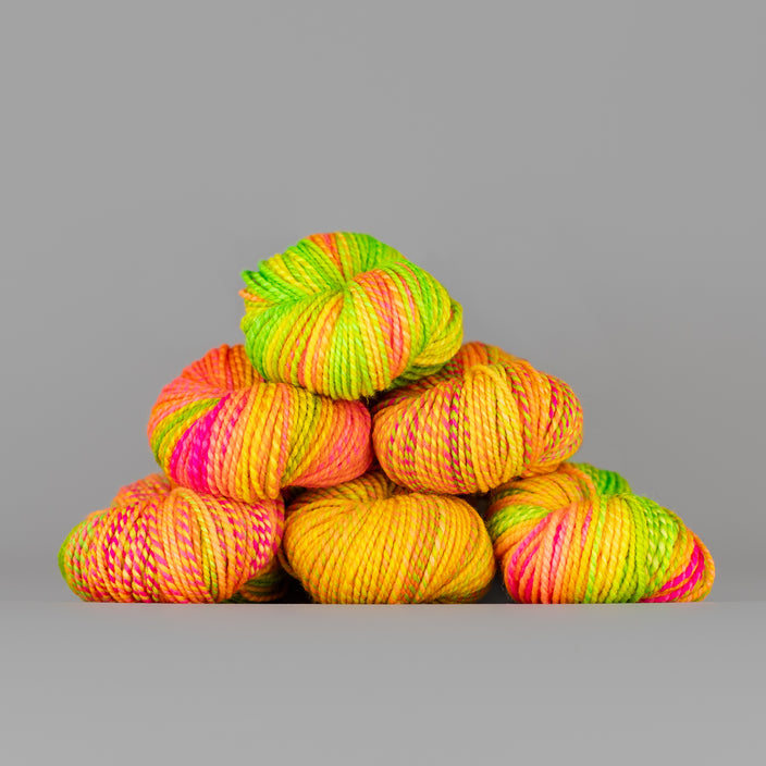 Skeins of Spincycle Dyed in the Wool sport weight yarn in a neon green, pink, orange, and yellow color gradient against a gray background.