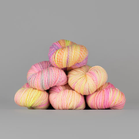Skeins of Spincycle Dyed in the Wool sport weight yarn in a pale yellow, pink, orange, and gray color gradient against a gray background.